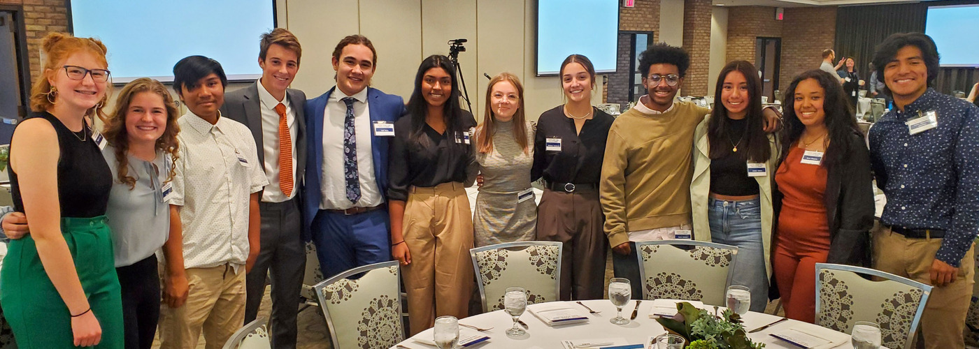 A group of 12 students standing with their arms around each other, smiling at the camera.