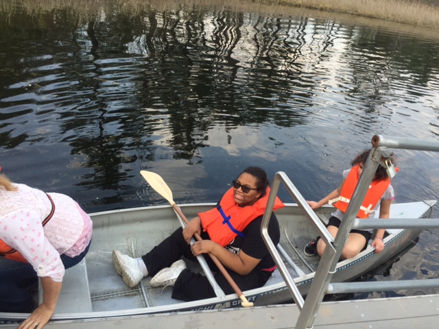 three students in a canoe on a lake, the middle student is looking up at the camera with an oar laying across her lap.