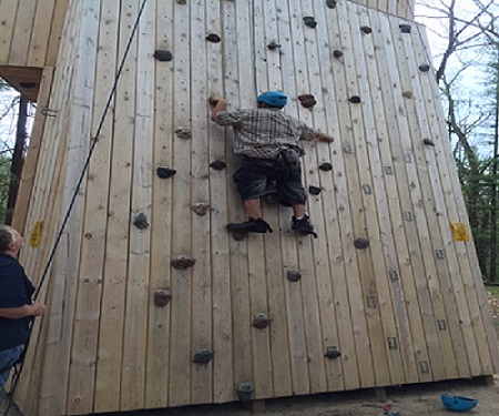 Young man climbing rock wall wearing a helmet, and harness. A man is on standing to the left, holding a rope that is connected to the harness holding up the student.